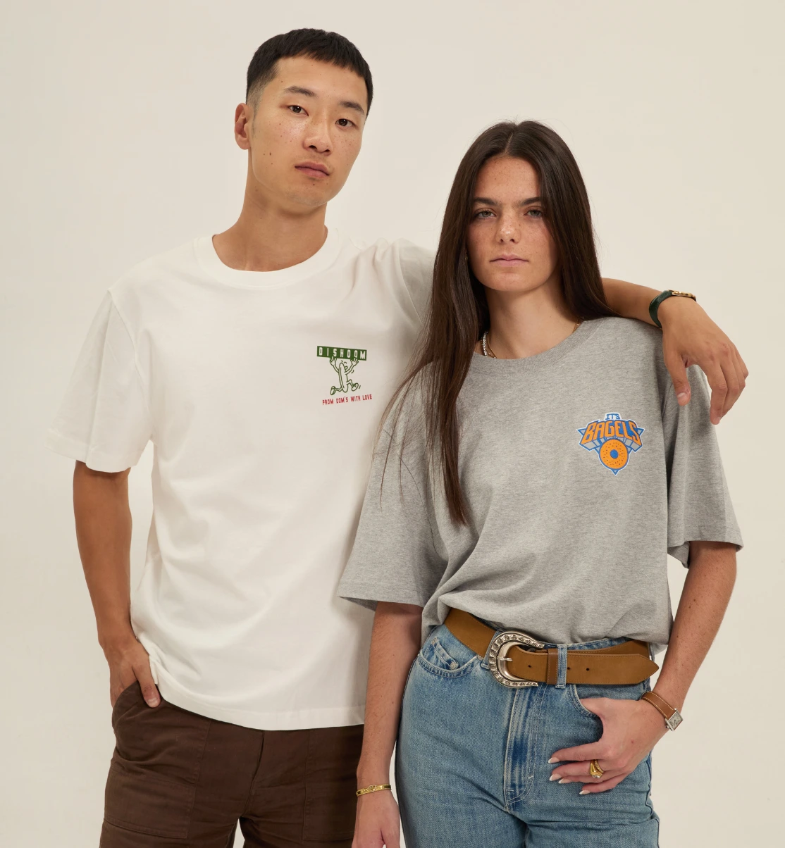 Lifestyle shot of premium branded apparel; featuring a man in a Dishoom logo tee and a woman in a grey heavyweight It's Bagels t-shirt with a relaxed fit